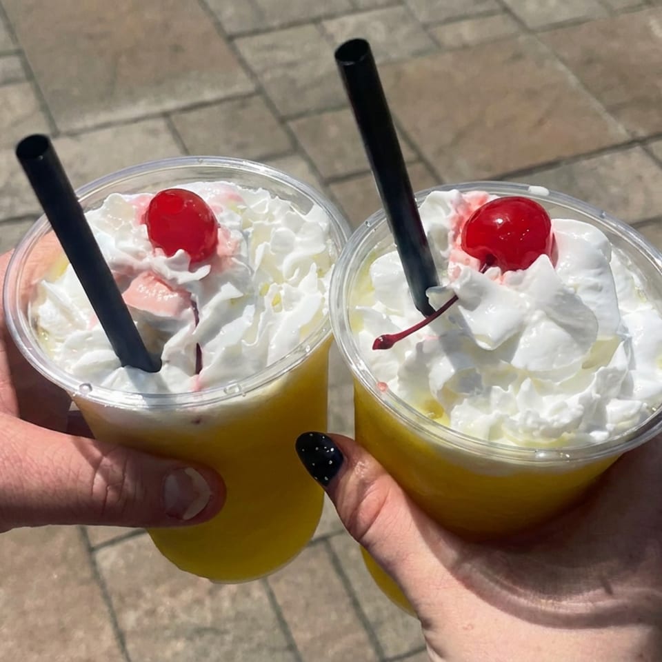 Two yellow whipped-cream drinks topped with cherries and black straws, held outdoors over a paved walkway.